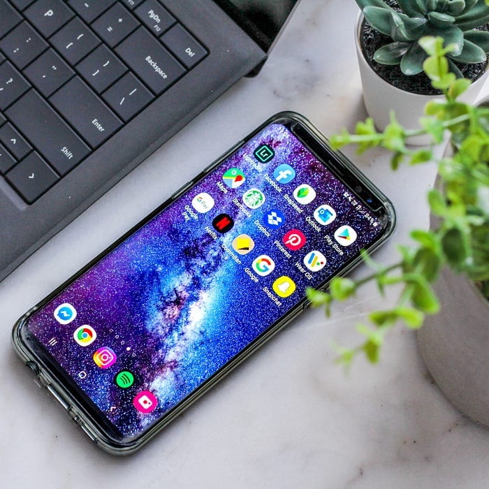 A smartphone with a vibrant space-themed wallpaper rests on a white surface. Its screen displays various app icons, including social media, streaming, and communication apps. The phone is next to a black laptop keyboard and surrounded by small potted plants.