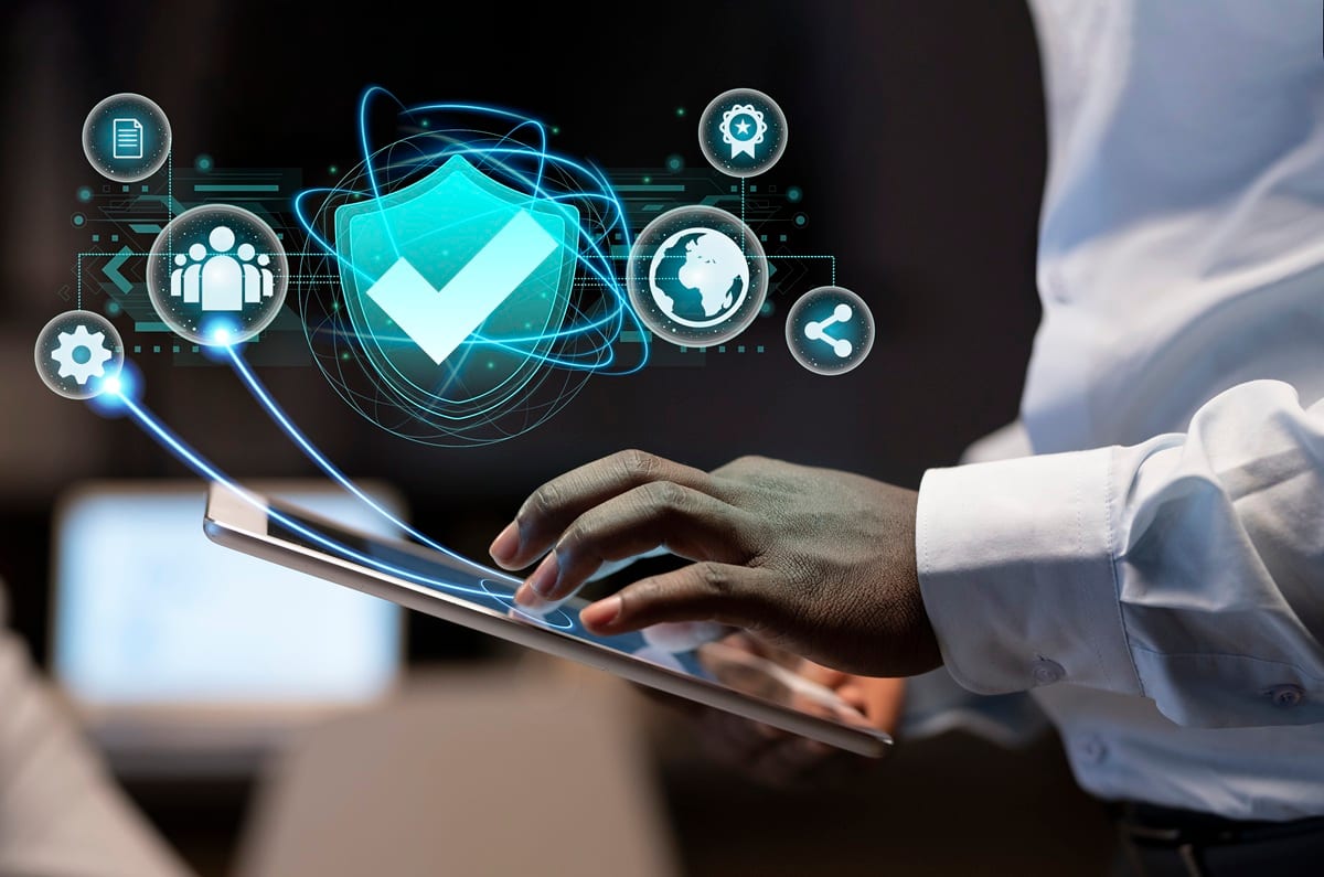 A person operates a tablet displaying a prominent digital shield icon surrounded by other connected symbols, visually representing cybersecurity, data protection, or network security management.