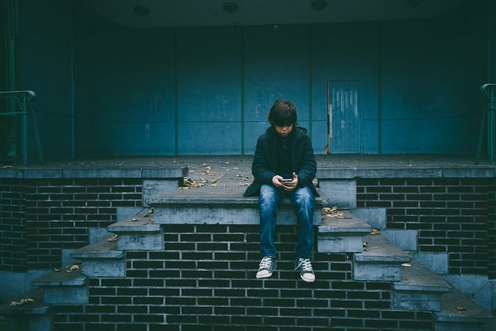 A child wearing a dark jacket and jeans sits alone on brick steps, looking at a handheld device. The background features a teal painted structure and scattered leaves. The scene has a moody and introspective atmosphere.