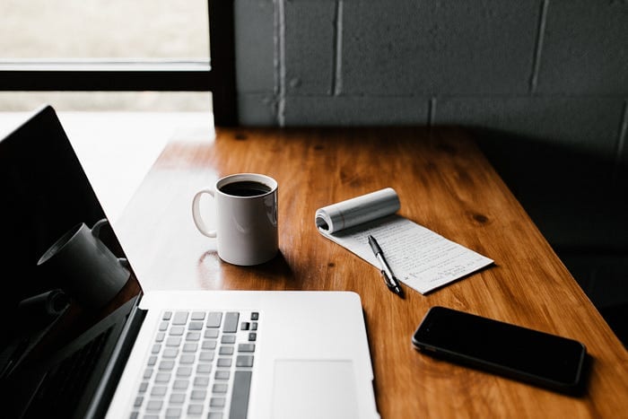 A wooden desk with a laptop, a white coffee mug, a notepad with written notes, a pen, and a smartphone. The desk is next to a window, allowing natural light to illuminate the workspace.