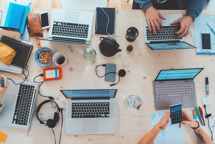 A wooden table is cluttered with laptops, notebooks, smartphones, headphones, a camera, a teapot, and various office supplies. Four people, only their hands visible, are typing on laptops, taking notes, and using smartphones. It appears to be a collaborative workspace.